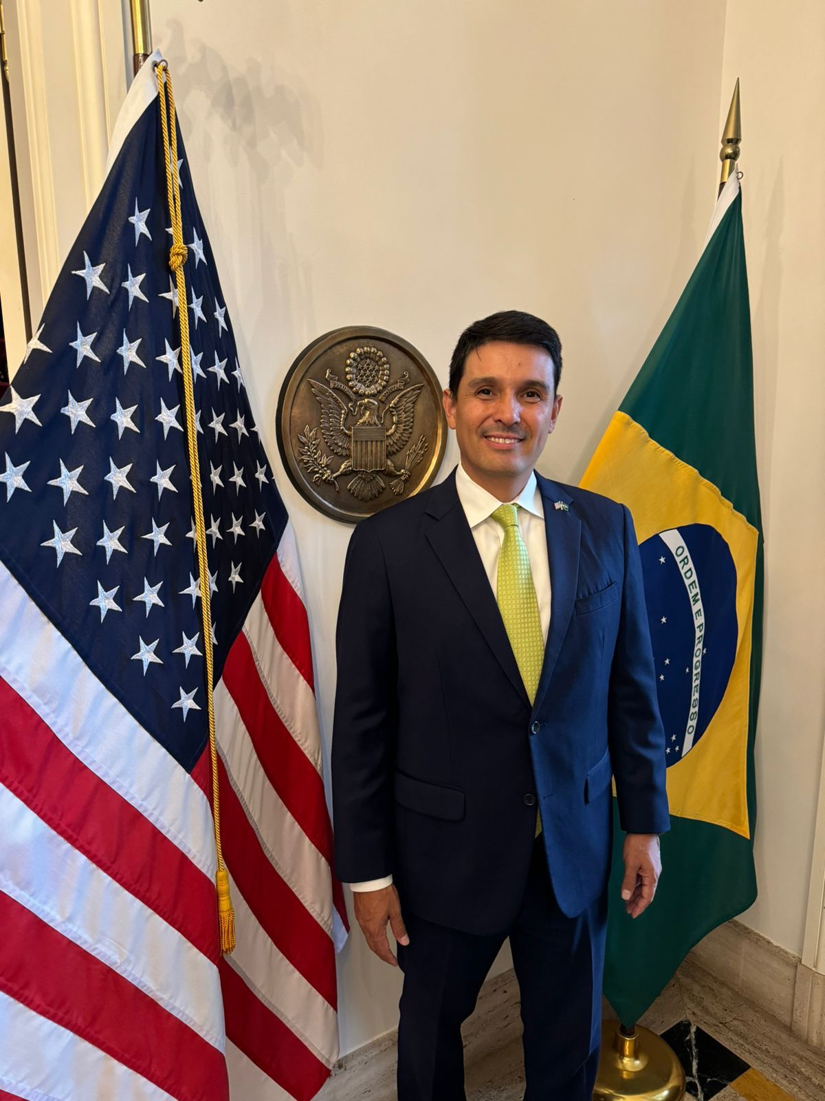 Cristiano Koga, CEO da MODERN Logistics, posa ao lado das bandeiras dos Estados Unidos e Brasil, em um ambiente formal, com o emblema dos EUA ao fundo.