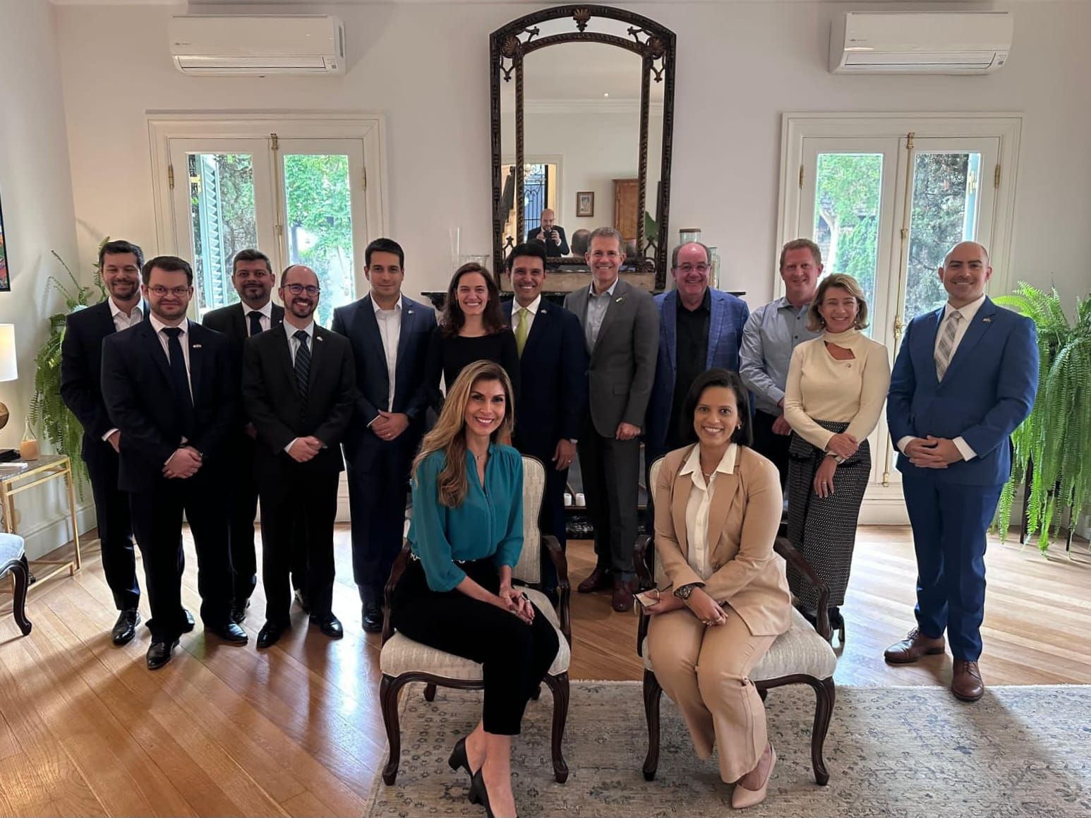 Um grupo de executivos de diversas empresas posando ao lado do Cônsul Geral dos EUA em São Paulo, em um ambiente elegante com janelas amplas e móveis clássicos.